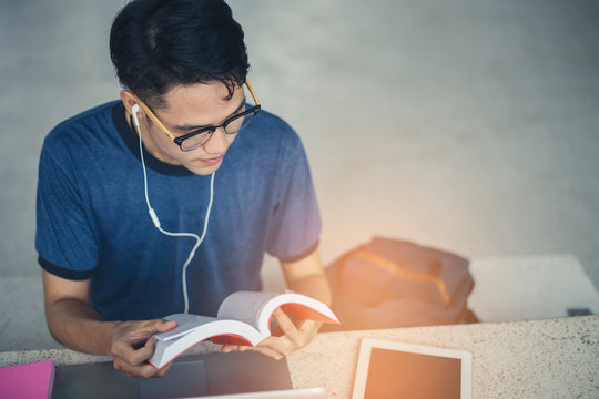 College Student Boy Reading Book For Exam And Listening To Music Via Headphones With Laptop.