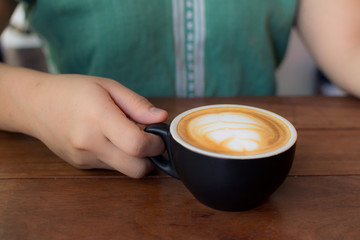 Latte Art, Coffe cup latte art, Hand of women holding a black cup of hot latte on wooden table. woman hand holding cup of coffee in the cafe, vintage tone.