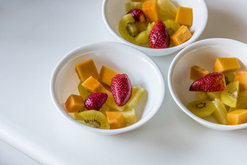 Colorful fresh fruit salad in white bowls, top view..Three white dishes of cut up healthy mixed fruit, white background, closeup..