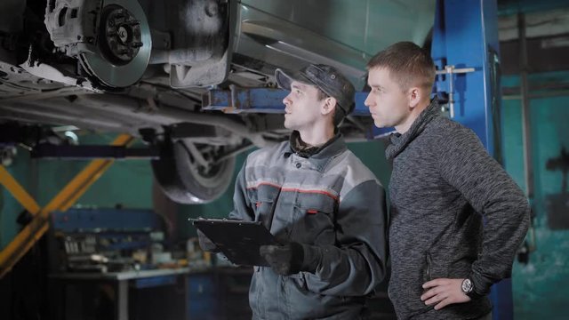 automobile mechanic is appraising a brake disk, brake pads and hosepipe, explaining for a car owner and calculating cost of repair, in a garage