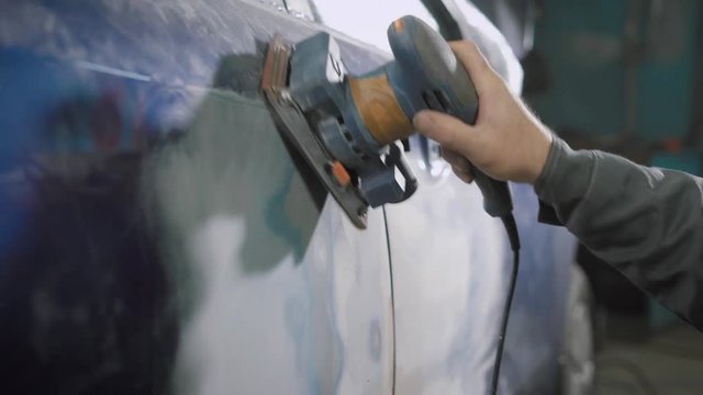 worker is brushing off old blue dye from automobile body using manual grinder, prepairing for painting in a car workshop