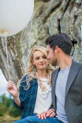 Portrait of the young beautiful smiling couple who standing outdoor and relying on old trunk of tree and holding the white balloon in hand