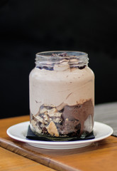In a glass jar a cake with different chocolate textures over a layer of crispy crumbled biscuits. Black background.