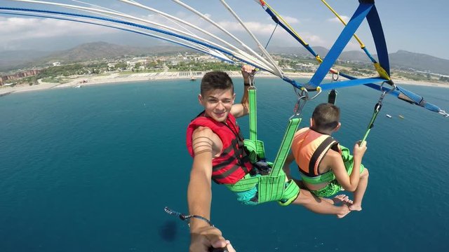 An amazing view of two boys flying over a Turkish resort on a multicolored parachute pulled by a motorboat making a selfie in slo-mo.   