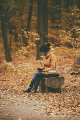 Young stylish hipster girl sitting on the bench in the autumn park and reading electronic book