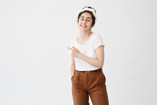 Advertising concept. Indoor shot of cheerful smiling young woman with curly hair in bun pointing index finger at copy space on white blank background