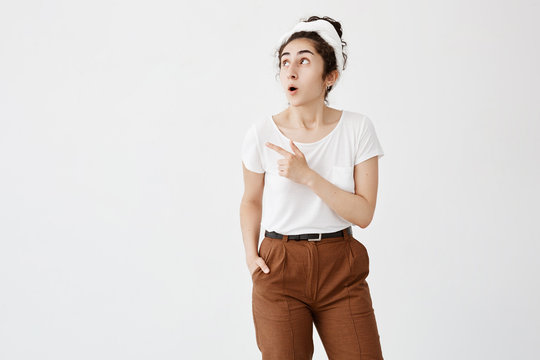Look at this! Cheerful young dark-haired woman with hair bun popped eyes in excitement, pointing her index finger away, indicating copy space on white blank wall for promotional information
