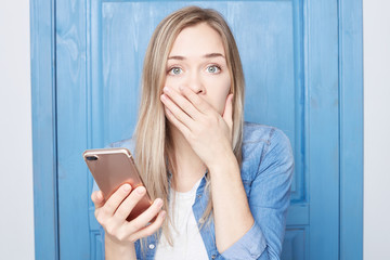 Fashionable stressful young female covers mouth in shock, got important message via sms, being very...