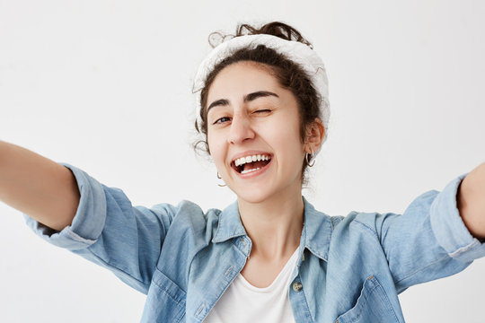 Happiness, joy, youth. Young positive girl dressed in denim shirt over white t-shirt stretching arms, smiling broadly, blinking, having good mood, demonstrating white even teeth