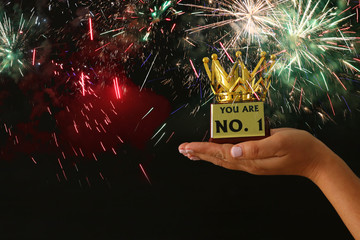 Woman's hand holding a crown trophy for show victory or winning first place over black background with glitter overlay.