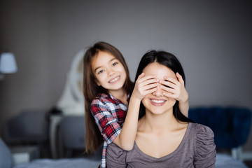 Beautiful young woman and her charming little daughter are hugging close mother eyes, looking at camera and smiling before make an surprise
