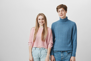 Cozy shot of Caucasian friends wearing knitted sweaters posing indoors. Hipster guy with fair hair and blue eyes standing behind his cute blonde girlfriend. Couple expressing positive emotions.
