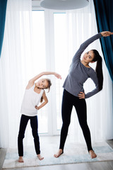 Beautiful young woman and charming little daughter are smiling while doing yoga together at home