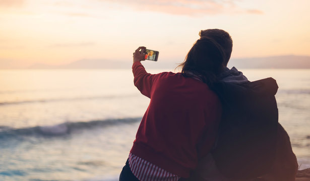 Couple hugging on background ocean sunrise, take photos on smartphone, two romantic people cuddling and looking on view evening seascape, hipster enjoy sunset together, love relax concept