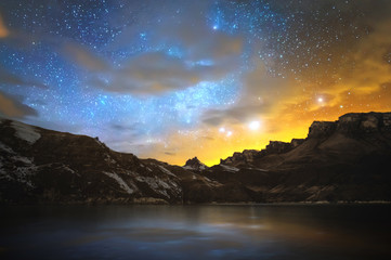 High mountain lake in the northern Caucasus, surrounded by epic rocks and a bright winter starry sky at sunset.