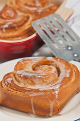   Close up freshly baked, homemade cinnamon buns with vanilla glaze drizzled on top, a metal spatula and red ceramic baking dish filled 