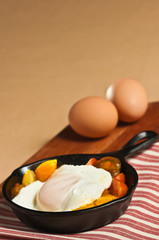 Freshly fried egg over light with sliced tomatoes in a hot, cast iron pan on a red and white stripped towel and two fresh brown eggs