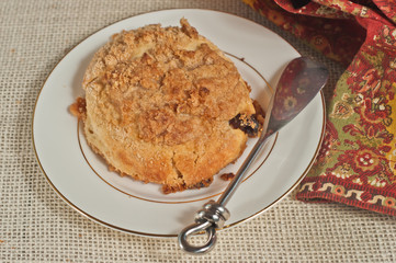 Close up of freshly baked, mini peach streusel coffee cakes on a white, round, plate with gold rim and an artisan spreading  knife with a print, cloth napkin on a tan burlap mat