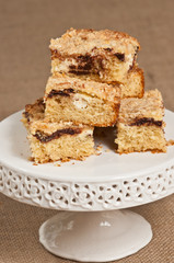 Close up of freshly baked, homemade, warm stack of ricotta and chocolate coffee cake squares on a round, white, pedestal display dish on a tan burlap mat