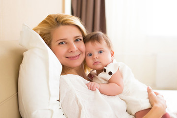 Happy loving family concept. Beautiful mother playing with her baby girl in the bedroom. They smiling and hugging together on white bed linens