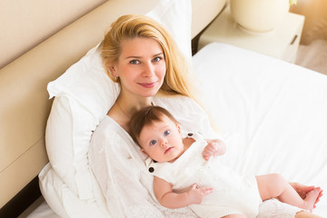 Obraz na płótnie Canvas Happy loving family concept. Beautiful mother playing with her baby girl in the bedroom. They smiling and hugging together on white bed linens
