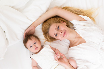 Obraz na płótnie Canvas Portrait of young mother with blond hair with her sweet 3 month old baby girl in white wear having fun in the bedroom at morning, loving happy family concept, top view