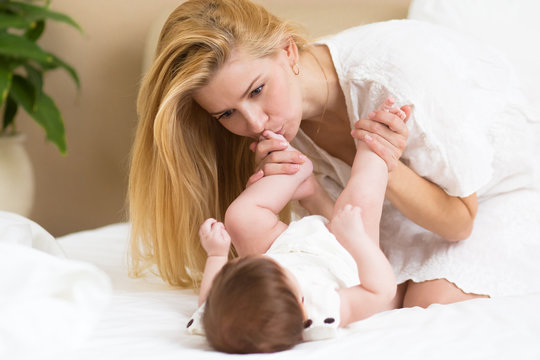 Mother and baby girl on a white bed. Mom and cute baby playing in sunny bedroom at home. Family having fun together. Young mother kissing her daugther feet