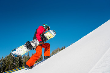 Full length shot of a fully equipped snowboarder walking up the slope at winter ski resort Bukovel, carrying his snowboard copyspace nature outdoors recreation seasonal sport active lifestyle concept