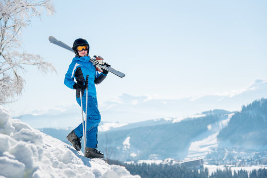 Full length shot of a female skier walking down the slope with her skis on the shoulder, smiling to the camera copyspace winter ski resort, blue sky and mountains on the background
