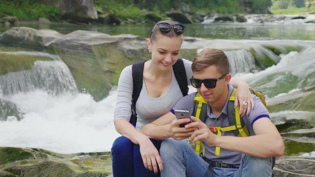 A young couple is sitting on a rock against a background of a rugged mountain river. They use a smartphone. Technology and travel concept