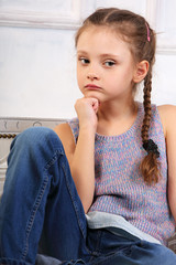 Fototapeta na wymiar Beautiful thinking cute kid girl sitting on the bench in blue jeans and looking sad. Closeup portrait