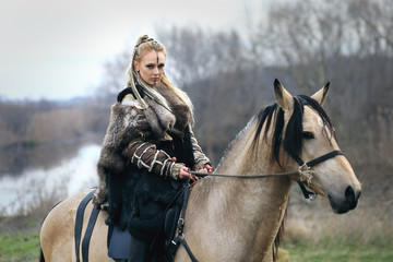 Beautiful viking warrior woman in traditional warrior clothes riding a horse in forest - Viking woman with makeup and brided hair riding horse in nature - 187141042