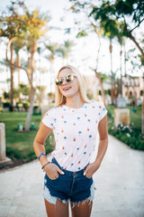 Fototapeta premium Attractive blonde woman in stylish sunglasses posing outside with exotic palm trees on background. Portrait of young woman with cute hairstyle walking down the street