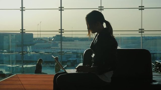 Young woman working with laptop in airport terminal. Waiting for my flight. Silhouette against the background of a large window