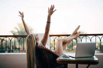 young woman with raised hands relaxing with laptop computer on a balcony on summer vocation