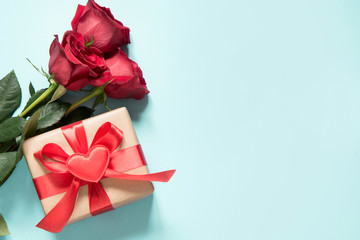 Bouquet of red roses and gift with red ribbon and heart on blue. Valentine's card. Close up. Copy space.