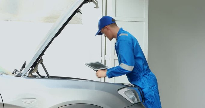 Male Caucasian mechanic checking a car machine while holding a digital tablet and wearing blue uniform, shot in 4k resolution