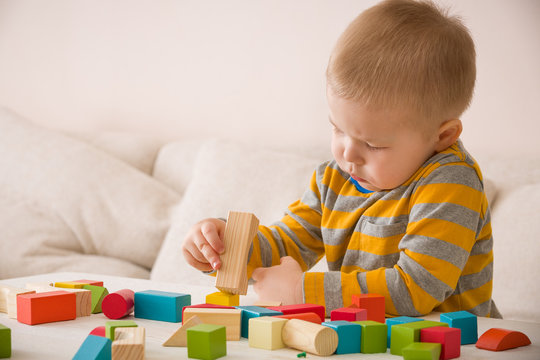Cute little toddler boy playing with colorful wooden bricks on the white table at home. Child building tower with geometric shapes. Learning and education concept.