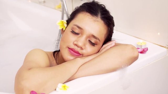Beautiful young woman relaxing in the bathtub and smiling at the camera with frangipani flowers in the bathroom
