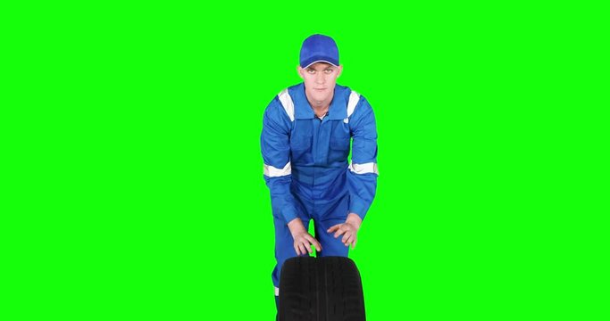 Caucasian auto mechanic pushing a tire in the studio while wearing blue uniform, shot in 4k resolution with green screen background