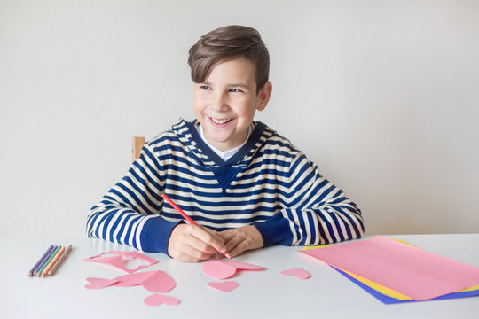 Valentine's Day. Mothers Day. Women's Day. Father's day. A tenage boy writes a valentine card for Valentine's Day. Sits at a white desk indoors. A pink heart made of paper. Pensive and beautiful smile