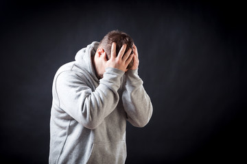 A young dark-haired man in a sporting gray sweatshirt is upset, upset and keeps his hands on his head on a black isolated background, the torturer suffers a headache