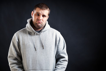 A young dark-haired man in a sports gray sweatshirt was upset that he fell ill and looks sad, his hands are holding in the pockets on a black isolated background