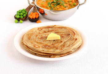 Kerala paratha, a type of Indian bread, with butter topping, and in the background is a wok of peas and carrot curry.