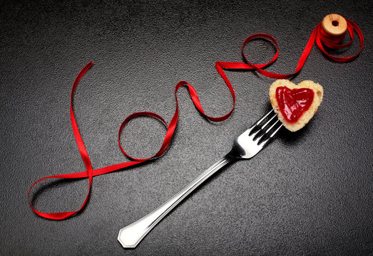 Inscription, word love of red satin ribbon and heart of toast bread with red jam on fork.Valentine day background.Love concept.On dark stone background.Creative.Love background