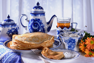 Freshly baked pancakes, a teapot, a glass with tea, sour cream are on a table set for the maslenitsa, pancake week celebration