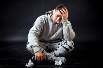 A young sad man in sports clothes got sick with flu and does not know how he is treated, put a napkin in his nose to stop the snot, put his hand on his head on a black isolated background.