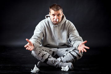 A young sad man in sports clothes got sick with flu and does not know how he is treated, stuck a napkin in his nose to stop the snot on a black isolated background.