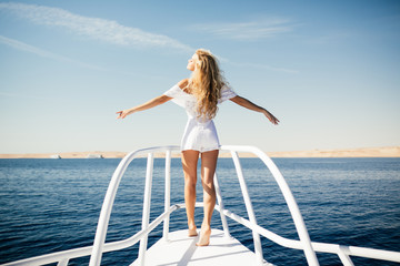 Woman standing on the nose of the yacht at a sunny summer day, breeze developing hair with open arms beautiful sea on background