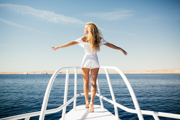 Woman standing on the nose of the yacht at a sunny summer day, breeze developing hair with open...